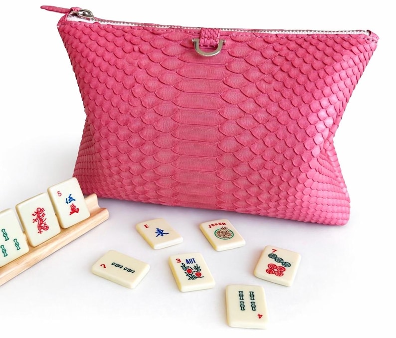 May include: A vibrant pink clutch with a textured snakeskin pattern, a silver zipper, and a small silver ring detail. Several ivory Mahjong tiles with colorful symbols and numbers are scattered around the clutch, and some are in a wooden rack.