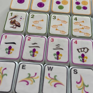 Puede incluir: Primer plano de fichas de Mahjong blancas con varios símbolos y números. Las fichas presentan imágenes de un pelícano, trompetas y una flor de lis. Algunas fichas muestran las letras E, W y S. También están presentes los números 1, 2, 3 y 4.