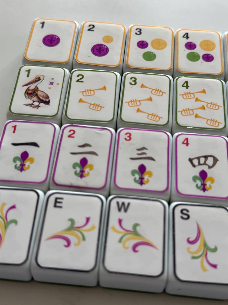May include: Close-up of white Mahjong tiles with various symbols and numbers. The tiles feature images of a pelican, trumpets, and a fleur-de-lis. Some tiles display the letters E, W, and S. The numbers 1, 2, 3, and 4 are also present.