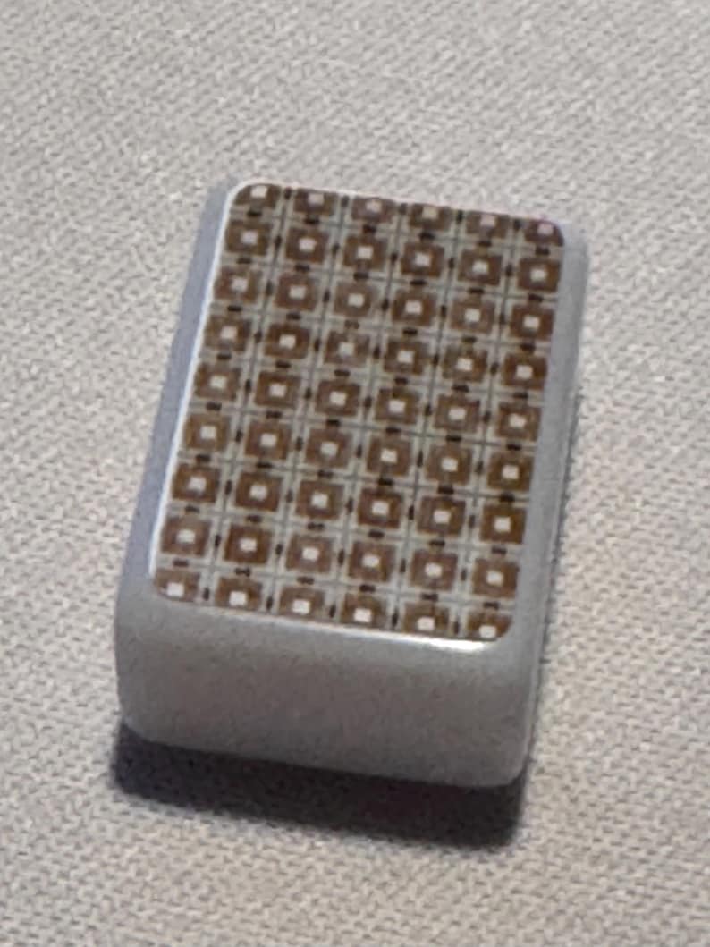 Op de afbeelding: Een rechthoekig, grijs object met een patroon. Het patroon bestaat uit bruine vierkanten met witte accenten, gerangschikt in een raster. Het object heeft afgeronde randen en lijkt van een glad materiaal te zijn gemaakt.