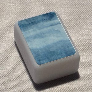 Op de afbeelding: Een rechthoekige, lichtgrijze gum met een blauw aquarelontwerp op het bovenvlak. Het blauwe ontwerp heeft een wolkachtig uiterlijk, met tinten licht- en donkerblauw. De gum heeft licht afgeronde randen.