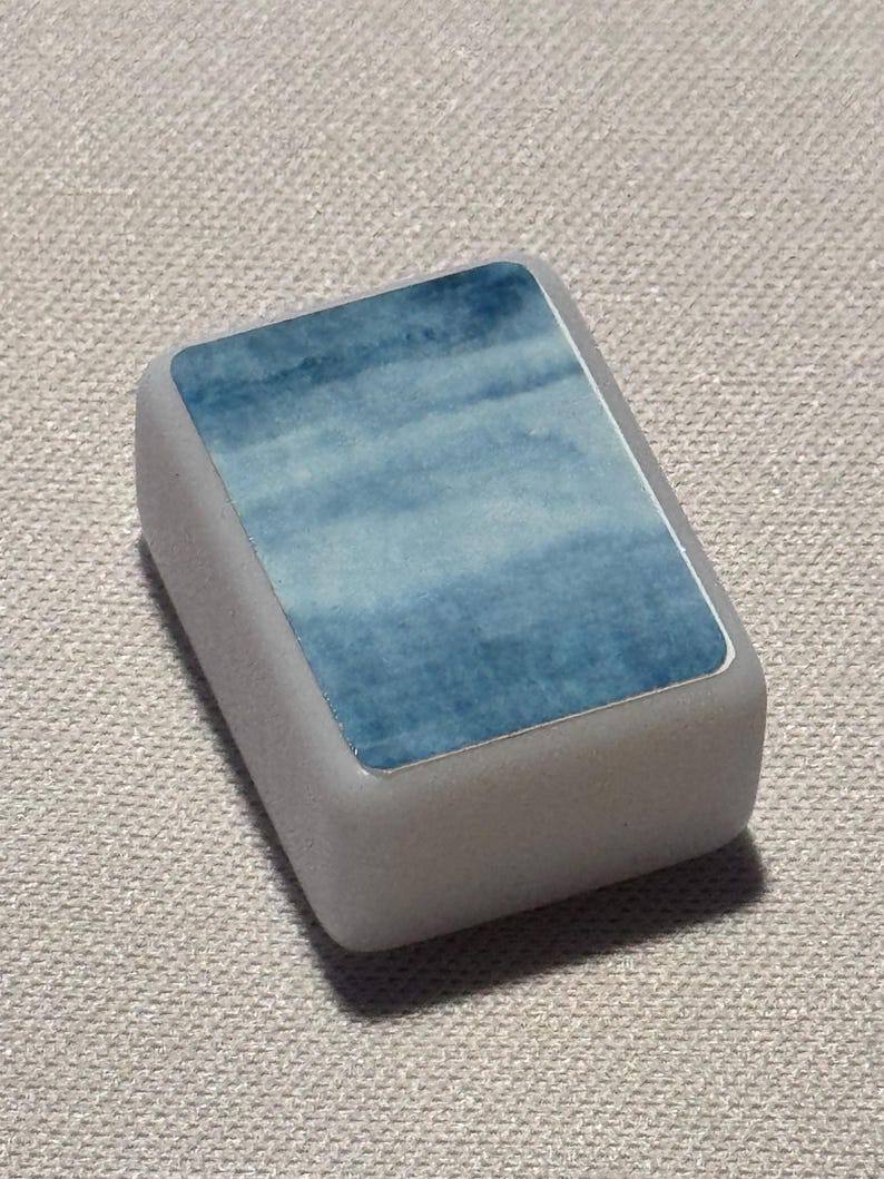 Op de afbeelding: Een rechthoekige, lichtgrijze gum met een blauw aquarelontwerp op het bovenvlak. Het blauwe ontwerp heeft een wolkachtig uiterlijk, met tinten licht- en donkerblauw. De gum heeft licht afgeronde randen.