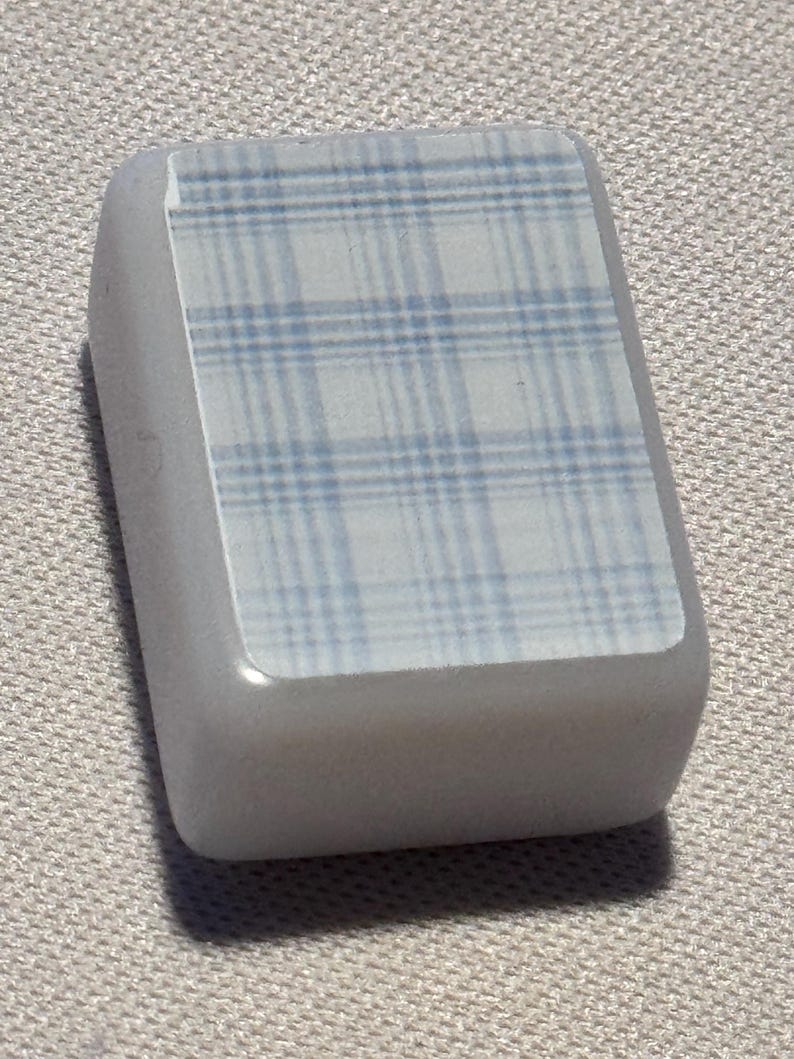 Op de afbeelding: Een lichtgrijs, rechthoekig object met een blauw-wit ruitpatroon op het bovenvlak. Het object heeft afgeronde randen en lijkt gemaakt van een glad, stevig materiaal. Het ruitpatroon heeft een raster van elkaar kruisende lijnen.