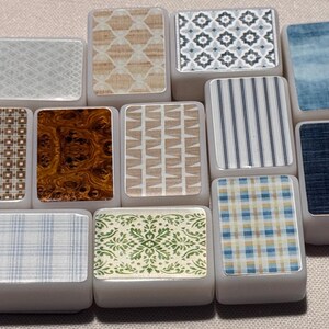 Op de afbeelding: Een verzameling rechthoekige tegels met diverse decoratieve patronen. Ontwerpen omvatten geometrische vormen, houtnerf, strepen en bloemmotieven. De tegels zijn in tinten blauw, bruin, beige en wit.