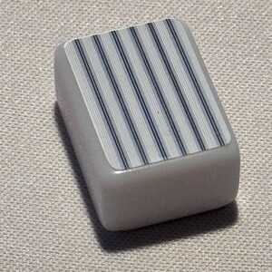 Op de afbeelding: Een rechthoekig, lichtgrijs object met een bovenoppervlak met een patroon van verticale blauwe en witte strepen. Het object heeft afgeronde randen en lijkt gemaakt te zijn van een glad, stevig materiaal.