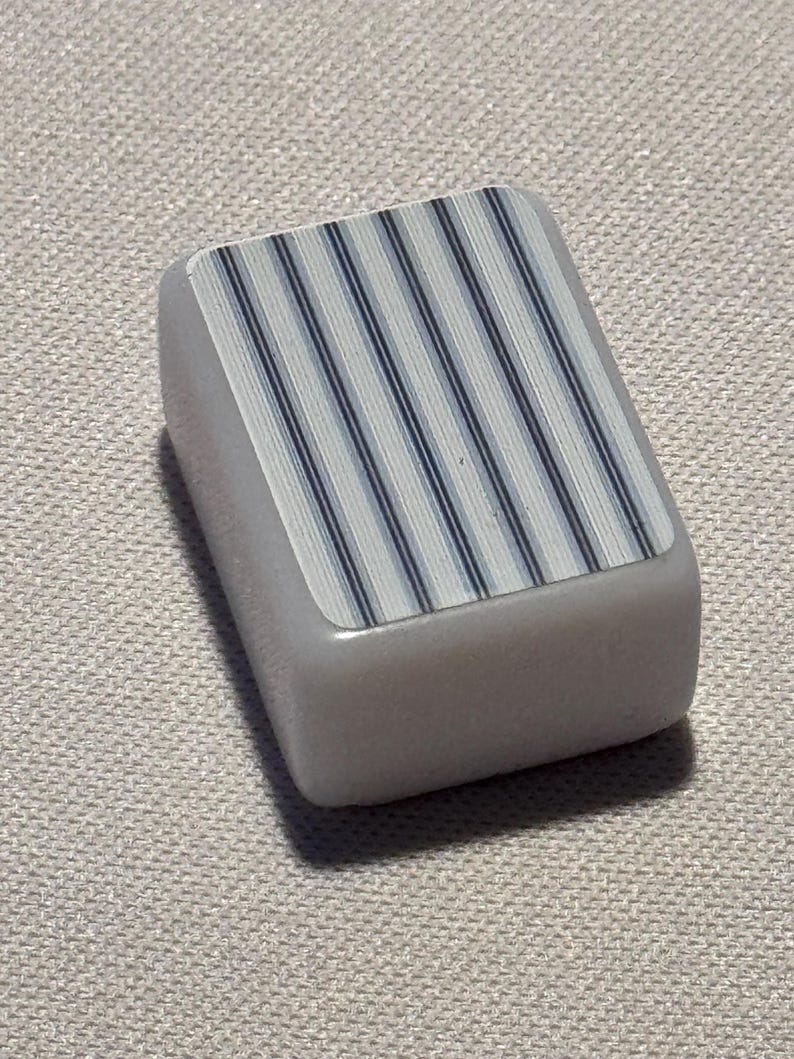 Op de afbeelding: Een rechthoekig, lichtgrijs object met een bovenoppervlak met een patroon van verticale blauwe en witte strepen. Het object heeft afgeronde randen en lijkt gemaakt te zijn van een glad, stevig materiaal.