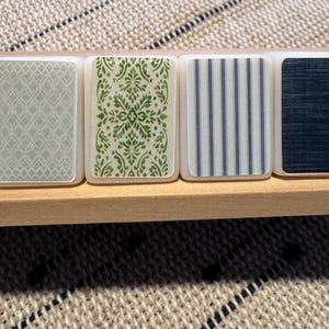 Op de afbeelding: Een close-up van decoratieve tegels met verschillende patronen. De tegels zijn rechthoekig met afgeronde hoeken en liggen op een houten plank. De patronen omvatten een grijs geometrisch ontwerp, een groen bloemenontwerp, blauwe en witte strepen en een donkerblauw textuurpatroon.