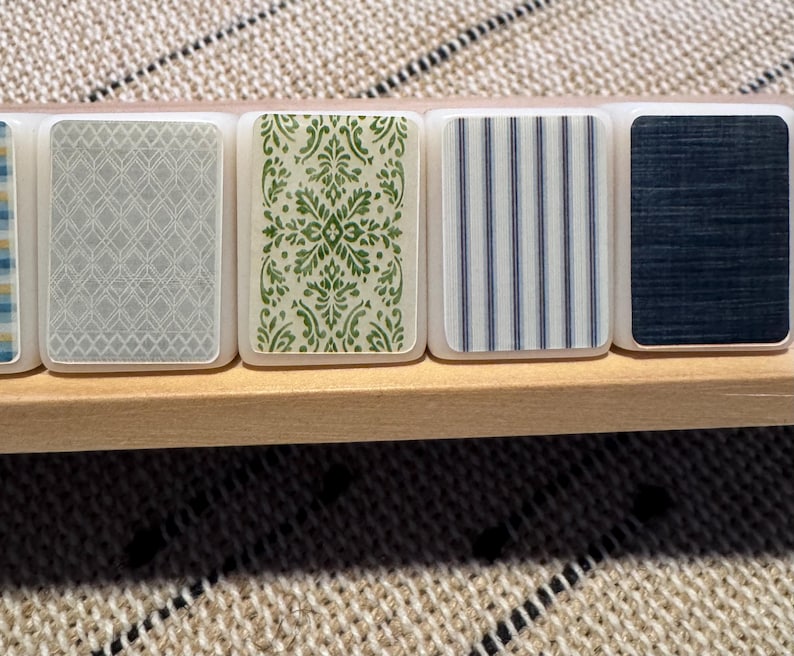 Op de afbeelding: Een close-up van decoratieve tegels met verschillende patronen. De tegels zijn rechthoekig met afgeronde hoeken en liggen op een houten plank. De patronen omvatten een grijs geometrisch ontwerp, een groen bloemenontwerp, blauwe en witte strepen en een donkerblauw textuurpatroon.