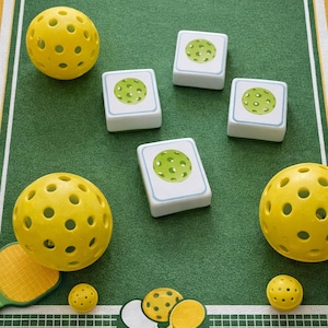 Op de afbeelding: Een pickleball-spelset met gele ballen, rackets en speelkaarten op een groene ondergrond. De set bevat geperforeerde gele ballen, kleine witte speelkaarten met een groen balontwerp en gele rackets.