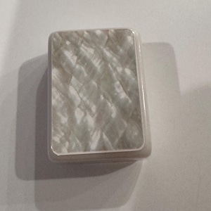 May include: A rectangular, iridescent, mother-of-pearl-like box with rounded edges. The surface has a textured, quilted pattern in shades of white and gray. The box is set against a plain white background.