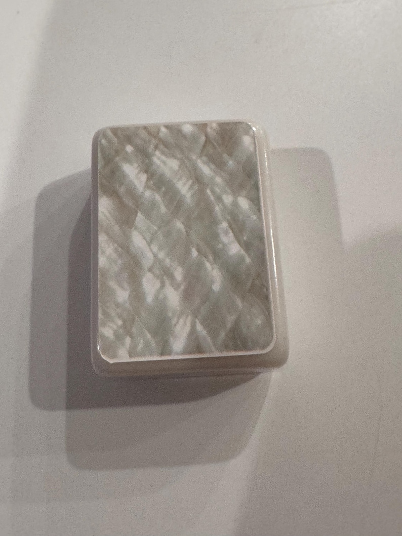 May include: A rectangular, iridescent, mother-of-pearl-like box with rounded edges. The surface has a textured, quilted pattern in shades of white and gray. The box is set against a plain white background.