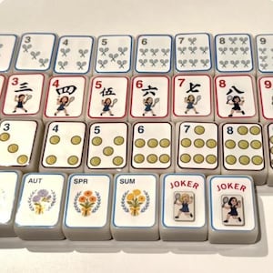 Peut inclure: Un ensemble de tuiles de Mahjong, pr&eacute;sentant divers symboles, chiffres et caract&egrave;res. Les tuiles sont blanches avec des accents rouges, bleus et dor&eacute;s. Certaines tuiles affichent des joueurs de tennis, d'autres des motifs floraux et le mot "JOKER".