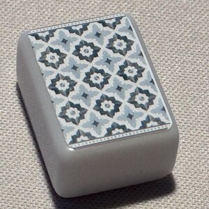 Op de afbeelding: Een rechthoekig, grijs object met een decoratieve bovenkant. De bovenkant heeft een herhalend bloemmotief in blauw, grijs en wit. Het object heeft een gladde, afgeronde vorm en is waarschijnlijk een decoratief element.