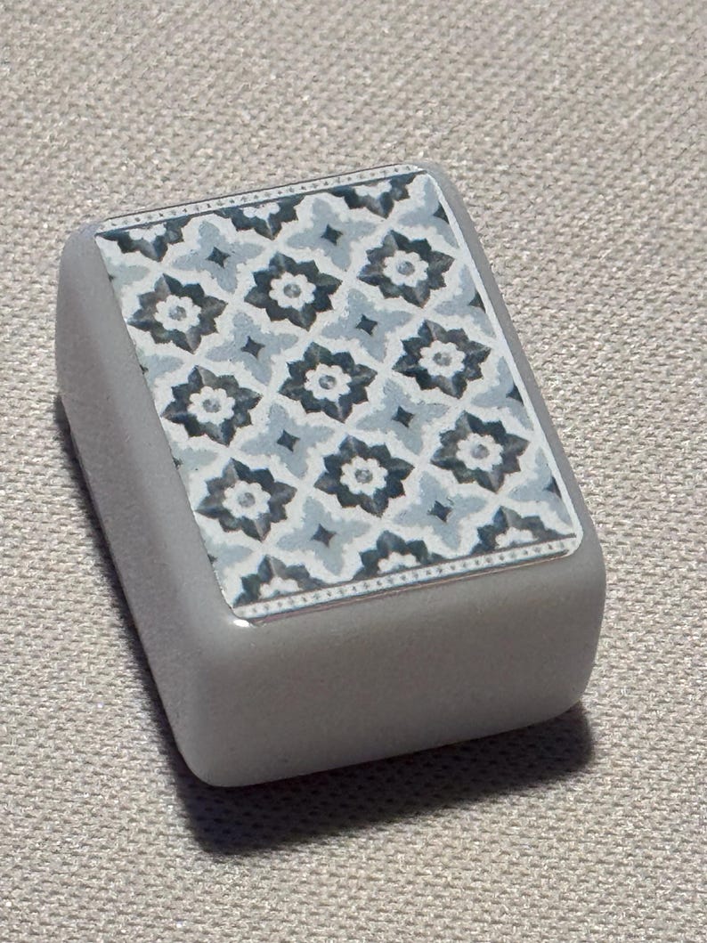 Op de afbeelding: Een rechthoekig, grijs object met een decoratieve bovenkant. De bovenkant heeft een herhalend bloemmotief in blauw, grijs en wit. Het object heeft een gladde, afgeronde vorm en is waarschijnlijk een decoratief element.
