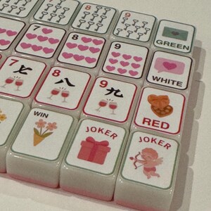 K&ouml;nnte beinhalten: Nahaufnahme eines Mahjong-Spielsteinesets. Die wei&szlig;en Spielsteine haben rosa R&auml;nder und zeigen Symbole, Zahlen und chinesische Schriftzeichen. Einige Spielsteine zeigen Herzen, Weingl&auml;ser und das Wort "JOKER".