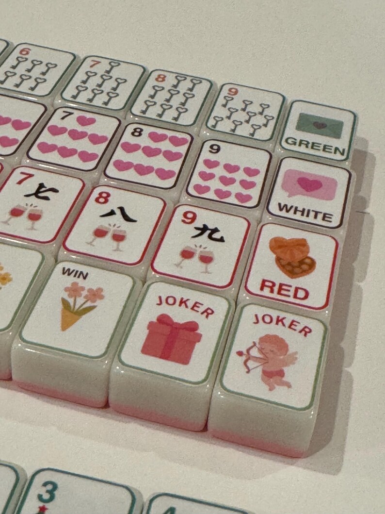 K&ouml;nnte beinhalten: Nahaufnahme eines Mahjong-Spielsteinesets. Die wei&szlig;en Spielsteine haben rosa R&auml;nder und zeigen Symbole, Zahlen und chinesische Schriftzeichen. Einige Spielsteine zeigen Herzen, Weingl&auml;ser und das Wort "JOKER".