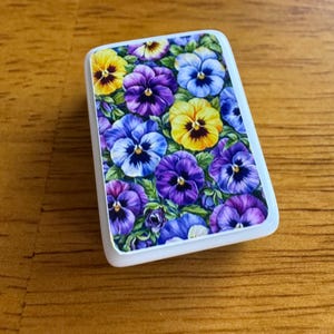 Peut inclure: Un objet d&eacute;coratif rectangulaire &agrave; bord blanc, orn&eacute; d'un motif floral de pens&eacute;es. Les pens&eacute;es pr&eacute;sentent des teintes de jaune, de violet et de bleu, sur un fond de feuilles vertes. L'objet est pos&eacute; sur une surface en bois.