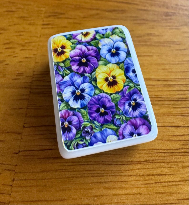 Peut inclure: Un objet d&eacute;coratif rectangulaire &agrave; bord blanc, orn&eacute; d'un motif floral de pens&eacute;es. Les pens&eacute;es pr&eacute;sentent des teintes de jaune, de violet et de bleu, sur un fond de feuilles vertes. L'objet est pos&eacute; sur une surface en bois.