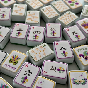 May include: A collection of white Mahjong tiles with purple, green, and yellow accents. The tiles feature various symbols, numbers, and Chinese characters. Some tiles display a fleur-de-lis design, while others have trumpet illustrations. The word "FLOWER" is printed on some tiles.