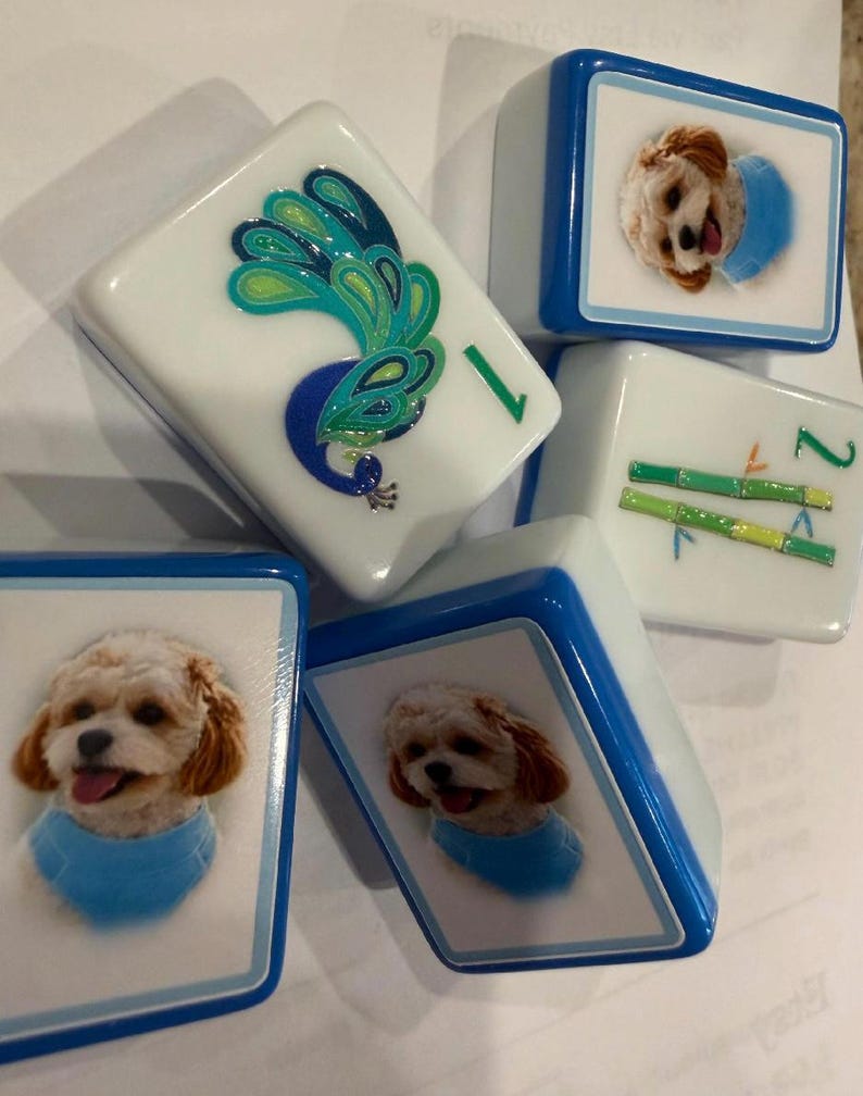May include: A collection of five rectangular mahjong tiles with white faces and blue edges. Two tiles feature a photographic portrait of a small light-coloured dog wearing a blue collar, one tile displays a green peacock, and one shows green bamboo stalks.