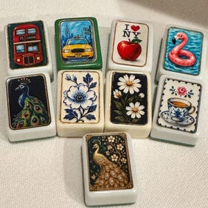 May include: A collection of nine rectangular mahjong tiles featuring diverse illustrations including a red London bus, a yellow taxi, an I heart NY apple, a pink flamingo float, a peacock, blue and white flowers, daisies, and a teacup.