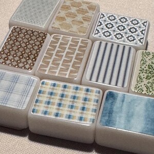 Op de afbeelding: Een verzameling rechthoekige tegels met verschillende patronen. De ontwerpen omvatten geometrische vormen, strepen en aquarelwashes in tinten blauw, bruin en beige. De tegels zijn gerangschikt op een neutraal gekleurd oppervlak.