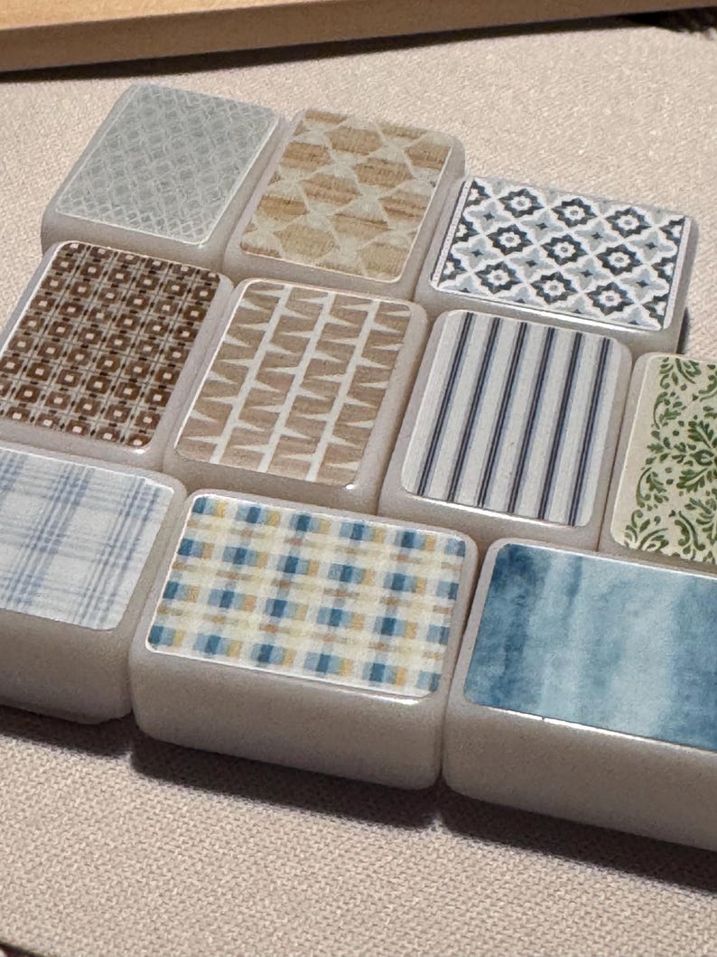 Op de afbeelding: Een verzameling rechthoekige tegels met verschillende patronen. De ontwerpen omvatten geometrische vormen, strepen en aquarelwashes in tinten blauw, bruin en beige. De tegels zijn gerangschikt op een neutraal gekleurd oppervlak.