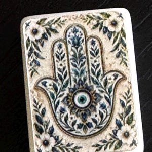 May include: A rectangular tile featuring a Hamsa hand symbol in the center, decorated with intricate blue and green floral patterns and a central eye motif. The background is a textured off-white, framed by small white flowers and green leaves.