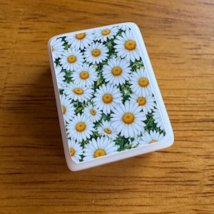 Peut inclure: Un objet rectangulaire blanc orn&eacute; d'un motif floral de marguerites &agrave; centres jaunes et feuilles vertes. L'objet a des coins arrondis et une finition brillante.