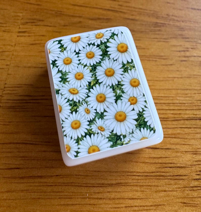 Peut inclure: Un objet rectangulaire blanc orn&eacute; d'un motif floral de marguerites &agrave; centres jaunes et feuilles vertes. L'objet a des coins arrondis et une finition brillante.