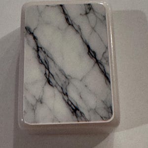 May include: A rectangular coaster with a marble design. The coaster features a white background with black veining, encased in a light-colored border. The design mimics the look of natural marble.
