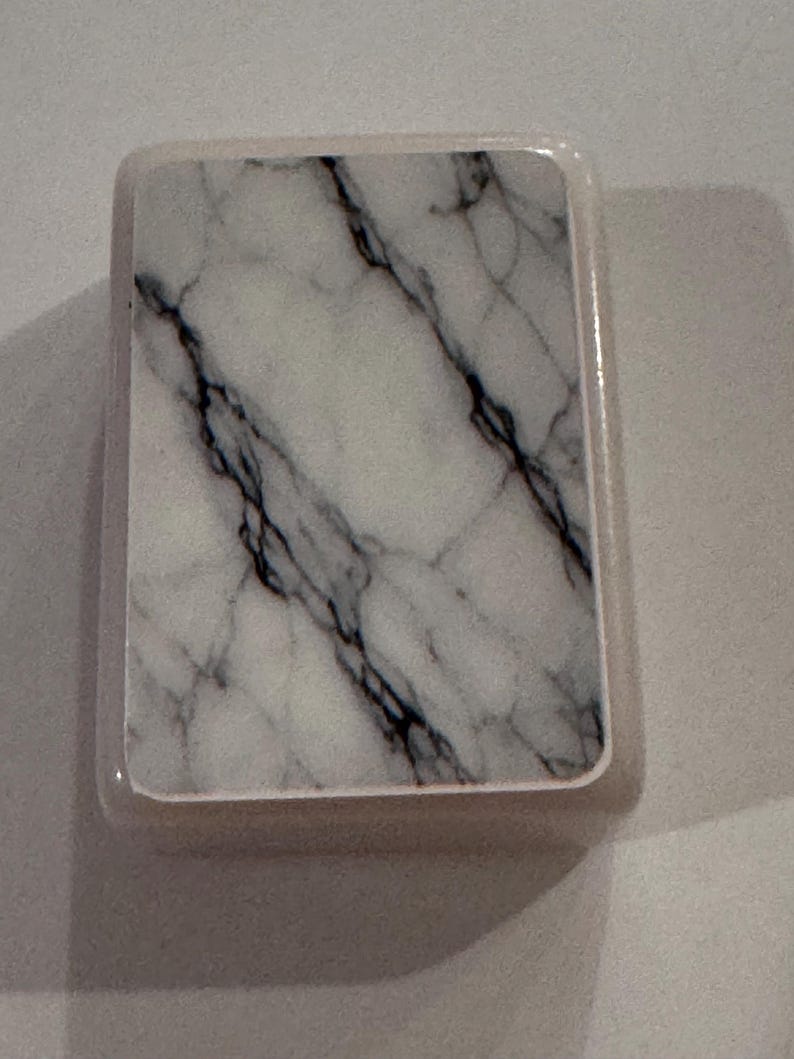 May include: A rectangular coaster with a marble design. The coaster features a white background with black veining, encased in a light-colored border. The design mimics the look of natural marble.