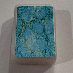 May include: A rectangular, light-colored stone with a turquoise and brown marbled design. The stone has rounded edges and a smooth surface. The turquoise pattern has a network of dark brown veins.