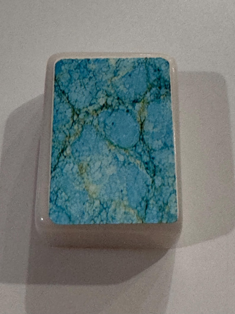 May include: A rectangular, light-colored stone with a turquoise and brown marbled design. The stone has rounded edges and a smooth surface. The turquoise pattern has a network of dark brown veins.