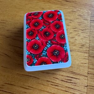 Peut inclure: Un objet rectangulaire bord&eacute; de blanc, orn&eacute; d'un motif floral. Le motif pr&eacute;sente de nombreux coquelicots rouges &agrave; centres noirs, parsem&eacute;s de petites fleurs bleues. L'aspect g&eacute;n&eacute;ral est vif et color&eacute;.
