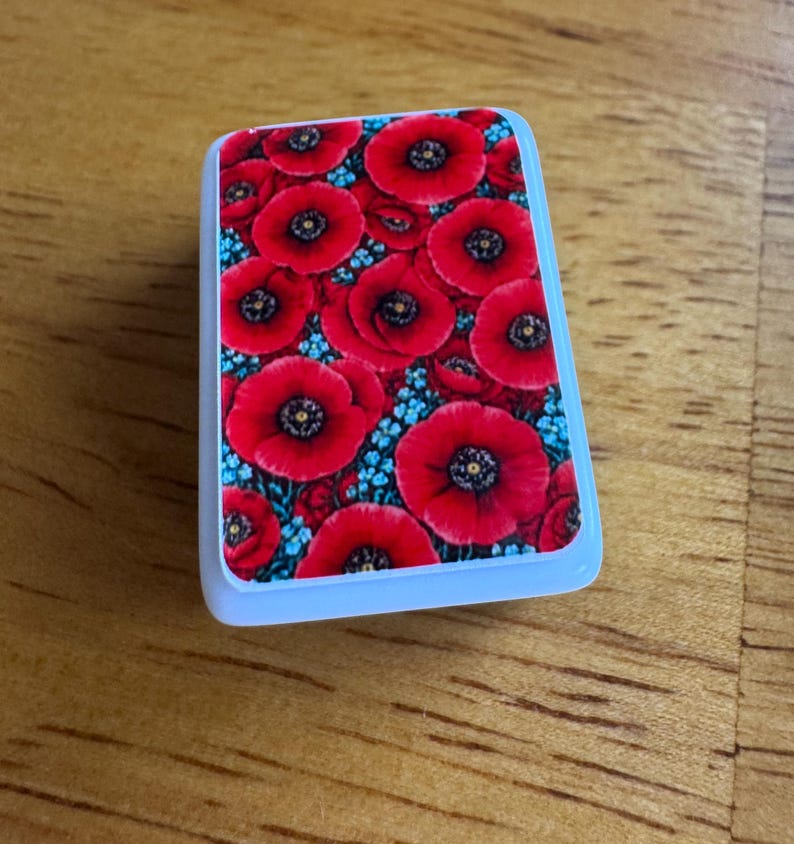 Peut inclure: Un objet rectangulaire bord&eacute; de blanc, orn&eacute; d'un motif floral. Le motif pr&eacute;sente de nombreux coquelicots rouges &agrave; centres noirs, parsem&eacute;s de petites fleurs bleues. L'aspect g&eacute;n&eacute;ral est vif et color&eacute;.