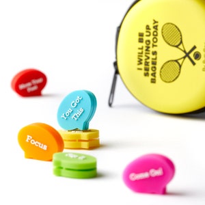 May include: A set of six colorful tennis ball holders with motivational phrases like "You Got This" and "Focus". The holders are shaped like speech bubbles and are designed to clip onto tennis balls. A yellow tennis ball case with the text "I Will Be Serving Up Bagels Today" is also shown.