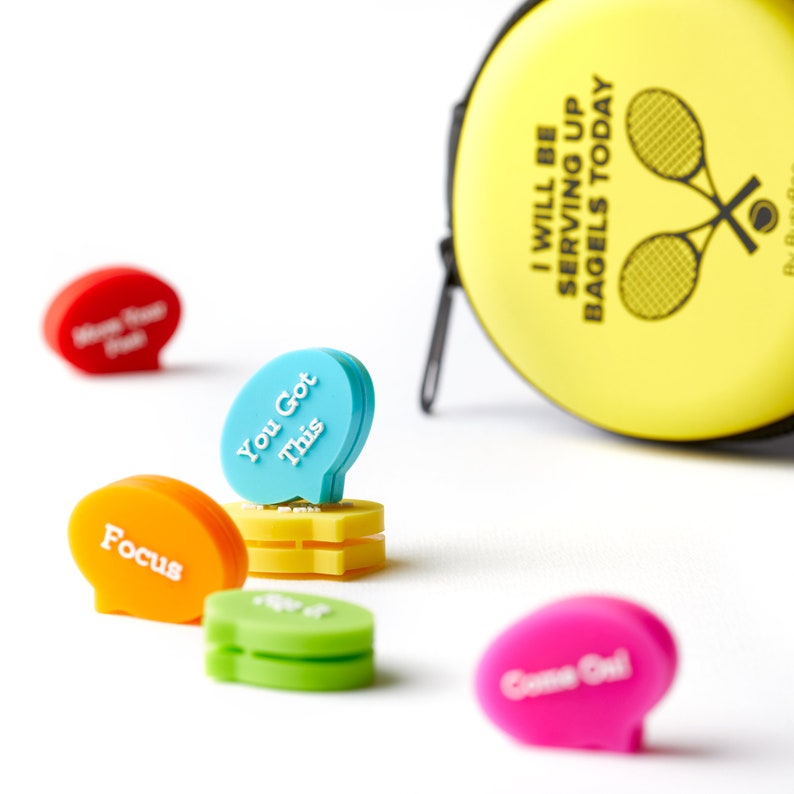 May include: A set of six colorful tennis ball holders with motivational phrases like "You Got This" and "Focus". The holders are shaped like speech bubbles and are designed to clip onto tennis balls. A yellow tennis ball case with the text "I Will Be Serving Up Bagels Today" is also shown.