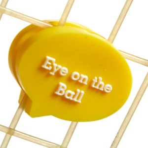 May include: Yellow plastic speech bubble with the text "Eye on the Ball" in white letters. The speech bubble is attached to a tennis racket.
