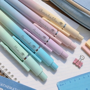 May include: A set of five pastel-colored retractable pens with clips, each personalized with a name and a graduation cap icon. The pens are arranged diagonally in shades of mint, blue, lavender, pink, and yellow, alongside a blue ruler.