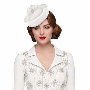 Pode incluir: Um chapéu pillbox branco com uma coroa arredondada e estruturada, apresentando um delicado laço de organza em camadas no topo. O chapéu é combinado com uma peça branca de manga comprida adornada com intrincados padrões florais de contas prateadas.