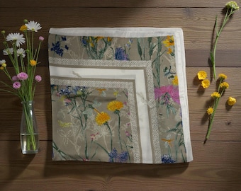 Könnte beinhalten: Ein quadratisches Tuch mit beigem Hintergrund und einem botanischen Druck aus gelben, blauen und rosafarbenen Wildblumen sowie grünen Stängeln. Das Tuch hat einen breiten weißen Rand mit einer feinen, gemusterten Borte, die das florale Zentrum abgrenzt.