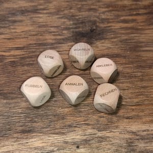 Könnte beinhalten: Sechs Würfel aus Holz mit deutschen Wörtern wie "Lose" und "Wurfel" sind auf einer Holzoberfläche verstreut. Die Würfel sind hellbraun mit dunkelbraunem Text. Der Hintergrund ist ein dunkelbrauner Holztisch.