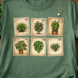 May include: Green t-shirt featuring a design of six postage stamps, each displaying a different houseplant illustration. The stamps have a vintage aesthetic with price markings. A gold necklace hangs on the shirt. The shirt is on a wooden surface.