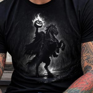May include: A black t-shirt featuring a graphic print of a headless rider on a horse in a dark forest. The rider holds a glowing, flaming jack-o'-lantern pumpkin head aloft. The design is rendered in shades of black, gray, and bright white.