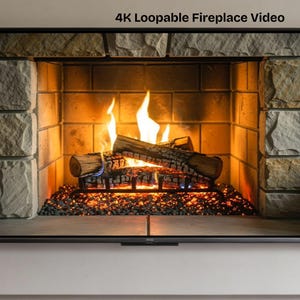 May include: A flat screen television displaying a 4K Loopable Fireplace Video. The screen shows a digital fireplace with a black metal grate, burning logs, glowing orange embers, and a surround made of light gray, textured rectangular stones.
