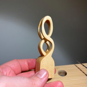 Op de afbeelding: Een handgesneden houten sculptuur met een abstract ontwerp. De sculptuur is gemaakt van lichtgekleurd hout en heeft een basis met een ontwerp dat lijkt op een oneindigheidssymbool. Het stuk wordt in de hand van een persoon gehouden.