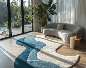 May include: A modern, wavy area rug in shades of blue and cream. The rug is placed on a light wood floor in a bright, airy room with a curved sofa and large windows. The text "Weavers Dream Rugs" is visible.