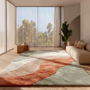 以下が含まれることがあります： 錆色、セージグリーン、クリーム色の抽象的なデザインの大きなラグ。 ソファ、木製のサイドテーブル、大きな窓のある部屋にラグが置かれています。 「Weavers Dream Rugs」の文字が見えます。