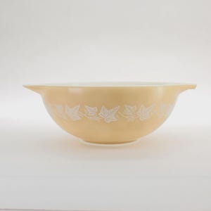 May include: A vintage, pale yellow mixing bowl with a white leaf and vine pattern around the exterior. The bowl has a rounded shape with a white interior and two small handles. A classic kitchenware item.